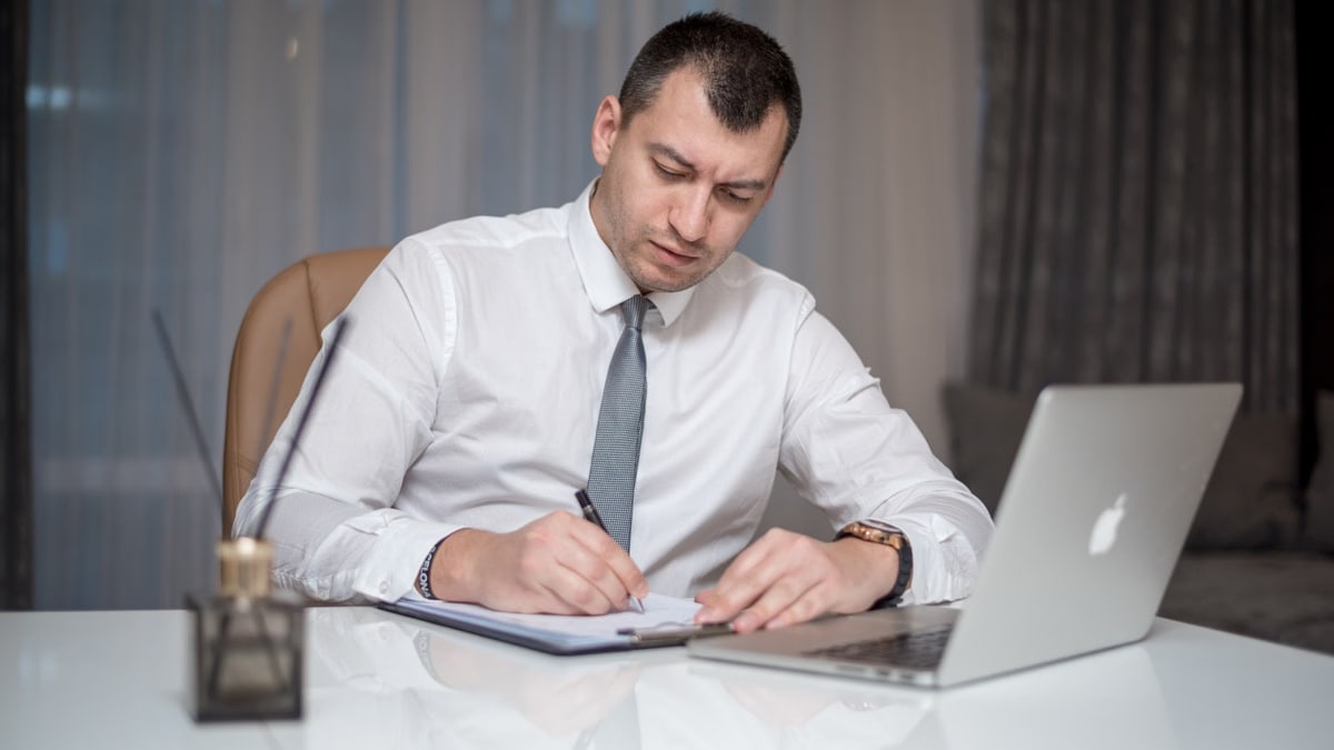 Businessman in suit writing notes at desk with laptop, reflecting modern office life.